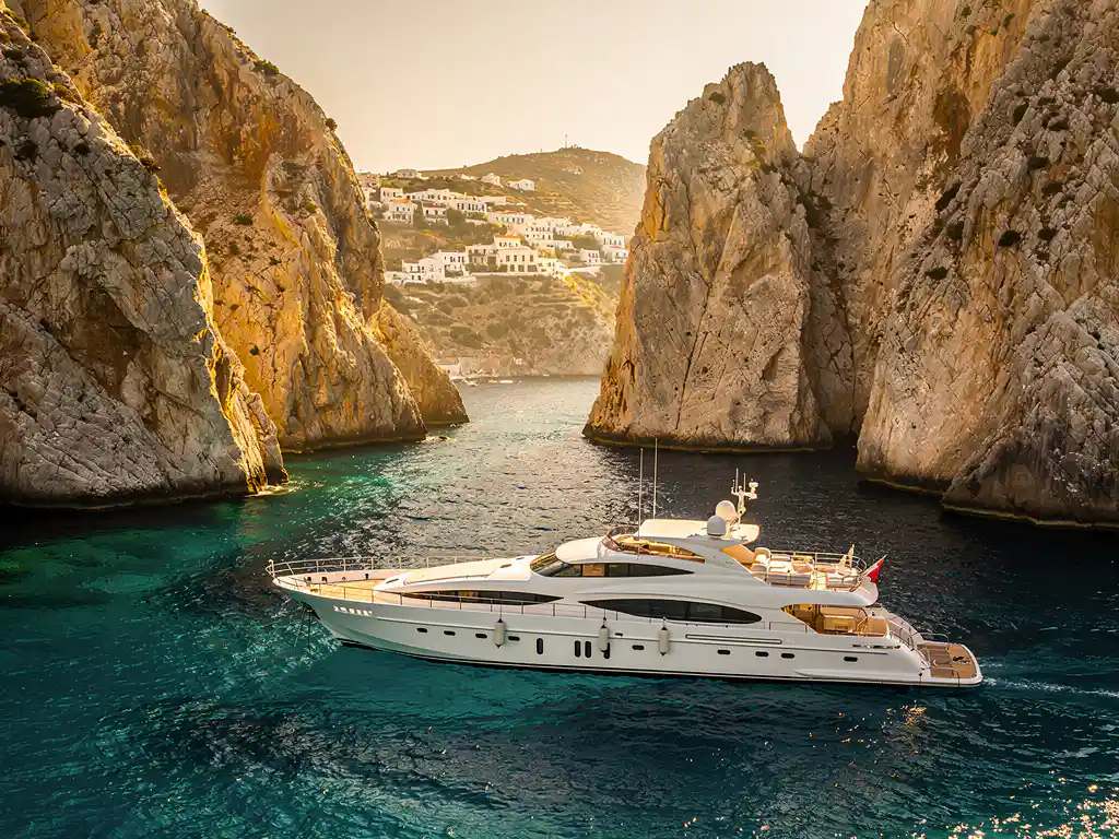 White luxury yacht anchored in turquoise Mediterranean cove between limestone cliffs with Greek village on distant hillside